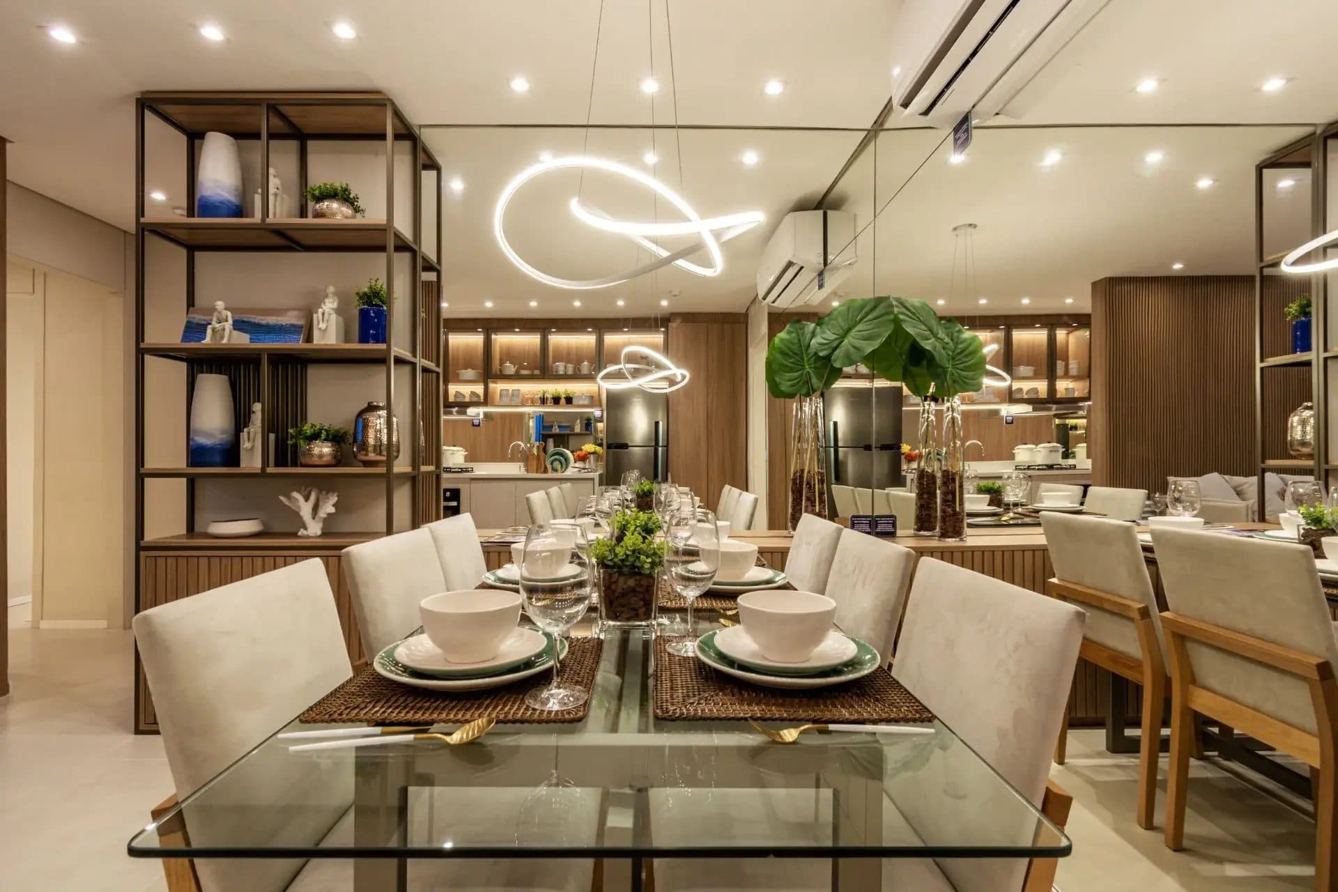 Sala de jantar com mesa de vidro, cadeiras estofadas, espelho na parede e iluminação moderna.